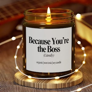Può includere: Una candela di soia accesa in un barattolo di vetro con la scritta "Because You're the Boss (Literally)". La candela è appoggiata su un sottobicchiere di legno ed è circondata da lucine. L'etichetta recita anche "organic | non toxic | soy candle".