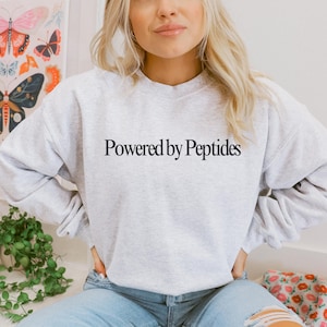 May include: A light gray sweatshirt with the text "Powered by Peptides" in black. The sweatshirt is a crew neck style and has long sleeves. The image shows a person wearing the sweatshirt.