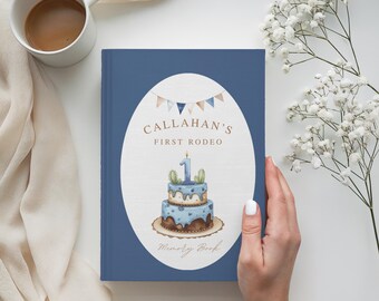 Puede incluir: Un libro de recuerdos azul con el texto "Callahan's First Rodeo" y una ilustración de pastel. El pastel tiene una vela con el número uno y decoraciones de cactus. El libro está sostenido por una mano, con una taza de café y flores en el fondo.