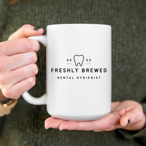 May include: A white ceramic mug featuring a black tooth graphic and the text "FRESHLY BREWED DENTAL HYGIENIST". The mug is held by a person wearing a green sweater.