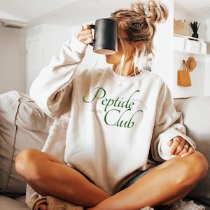 Peut inclure: Un sweat-shirt crème avec le texte "Peptide Club" en vert. Le sweat-shirt porte également le texte "EST 2024" et "make wellness" en vert. Une tasse noire est tenue devant le visage.