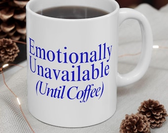 Puede incluir: Taza de cerámica blanca llena de líquido oscuro, con el texto azul "Emotionally Unavailable (Until Coffee)". La taza tiene un asa curva. Se ven piñas decorativas en el fondo.