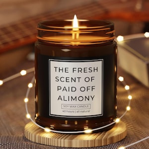 May include: A lit soy wax candle in a brown glass jar with a black label. The label reads "THE FRESH SCENT OF PAID OFF ALIMONY". The candle sits on a wooden coaster and is surrounded by fairy lights.