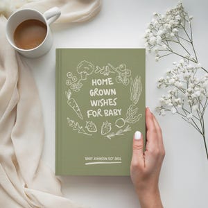 以下が含まれることがあります： 「Home Grown Wishes For Baby」の文字と野菜のイラストが入ったセージグリーンのベビー用ジャーナル。ジャーナルには「Baby Johnson Est 2026」という名前も含まれています。白いコーヒーカップと白い花が背景にあります。