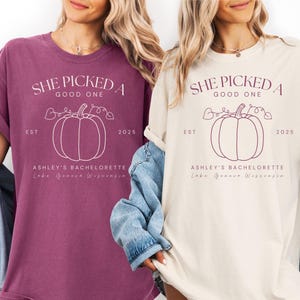 May include: Two t-shirts, one in a deep purple and the other in cream, each featuring a pumpkin graphic and the text "SHE PICKED A GOOD ONE". The text also includes "ASHLEY'S BACHELORETTE Lake Geneva Wisconsin" and the year "2025".