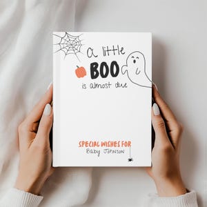 Könnte beinhalten: Ein weißes Buch mit dem Text "A little BOO is almost due" und "Special Wishes for Baby Johnson" in Orange und Schwarz. Das Cover zeigt einen Kürbis, ein Spinnennetz, einen Geist und eine Spinnenillustration.