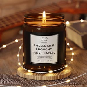 May include: A lit soy wax candle in a brown glass jar with a black label that reads "Smells Like I Bought More Fabric." The candle sits on a wooden coaster, surrounded by string lights. The candle is all natural and has a 40-hour burn time.
