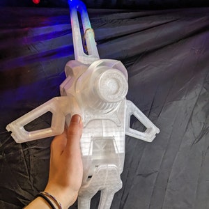 May include: A clear, 3D-printed object with a complex design, featuring a central cylindrical component and wing-like extensions. The object is held by a hand, showcasing its size and intricate details. The background is a dark, textured surface.