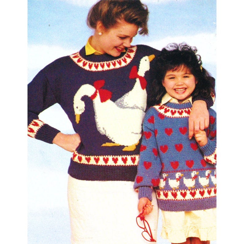 May include: Two blue knitted sweaters with a goose design. The larger sweater has two geese with red bows. The smaller sweater has a row of geese with red hearts. Both sweaters have a white heart border.