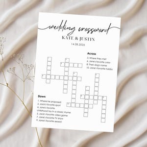 Wedding crossword puzzle card on white linen background with dried baby's breath flowers, minimalist black script "wedding crossword" header, couple names and personalized clues