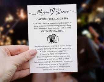 May include: A white card with black text and a camera icon. The card says "Megan Steven Capture the Love: I Spy" and lists a series of wedding-related moments to photograph, such as "Bride and groom sharing a joyous laugh" and "Selfie with a new friend!" The card also includes the hashtag #WEDDINGHASHTAG.