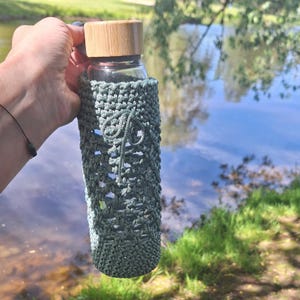 May include: A clear glass water bottle with a light brown wooden cap, encased in a sage green crocheted sleeve featuring an intricate openwork pattern and a decorative cord detail tied in a small loop on the front surface of the sleeve.