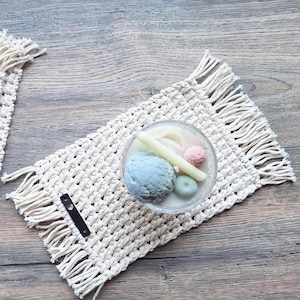 Könnte beinhalten: Ein cremefarbener Makramee-Untersetzer mit Fransen, der einen transparenten Glasbehälter mit einer Kerze und dekorativen Wachskugeln in Blau, Rosa und Grün hält. Die Kerze ist weiß. Der Untersetzer liegt auf einer Holzoberfläche.