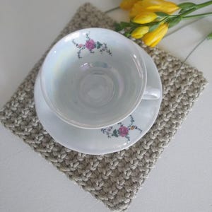 May include: A white ceramic teacup and saucer set with an iridescent finish, featuring a delicate floral pattern of pink roses and green leaves on the interior rim and saucer edge, resting on a square beige crocheted textile mat.