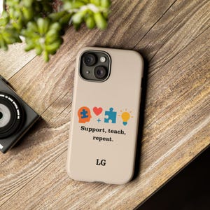 Könnte beinhalten: Beigefarbene Handyhülle mit einer bunten Grafik mit einem Puzzleteil, einem Herz und einer Glühbirne, mit dem Text "Support, teach, repeat". Die Initialen "LG" stehen unten. Die Handyhülle liegt auf einer Holzoberfläche.
