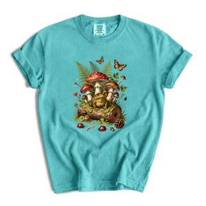 May include: A teal short-sleeve t-shirt featuring a graphic print of a toad sitting on a log surrounded by red-capped mushrooms, green ferns, orange butterflies, ladybugs, and a pinecone. The inner neck label displays the text Comfort Colors.
