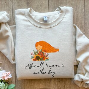 Puede incluir: Una sudadera de color beige claro con un gráfico de una mujer con cabello largo y rojo sosteniendo un girasol. El texto "After all, tomorrow is another day." está impreso debajo del gráfico.