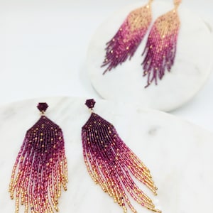 Può includere: Orecchini pendenti con perline bordeaux, rosa e oro. Gli orecchini presentano un effetto sfumato, che passa dal bordeaux scuro all'oro. Sono esposti su una superficie bianca.