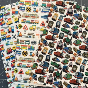 May include: Three fabric squares with different train-themed patterns. The first square has a white background with colorful trains, railroad crossing signs, and the words "Rail Crossing" and "Choo Choo". The second square has a white background with colorful trains, railroad crossing signs, and the words "Station" and "Rail Crossing". The third square has a white background with a repeating pattern of Thomas the Tank Engine and other characters from the Thomas & Friends franchise.
