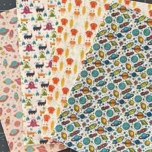 May include: Four different fabric sheets with various space-themed patterns. One sheet features a pink background with small planets and stars. Another sheet has a white background with colorful rockets and planets. The third sheet has a white background with orange and yellow aliens. The final sheet has a white background with green and purple aliens.