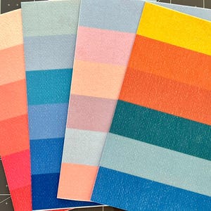 May include: A collection of colorful, textured paper rectangles. Each piece features horizontal stripes in varying shades of red, pink, blue, and yellow. The papers are arranged on a dark surface, showcasing their vibrant hues.