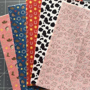 May include: Five different patterned fabric sheets. The sheets are all pink, red, blue, black and white, and white with a pink background. The patterns include cowboy hats, horseshoes, stars, and cow spots.