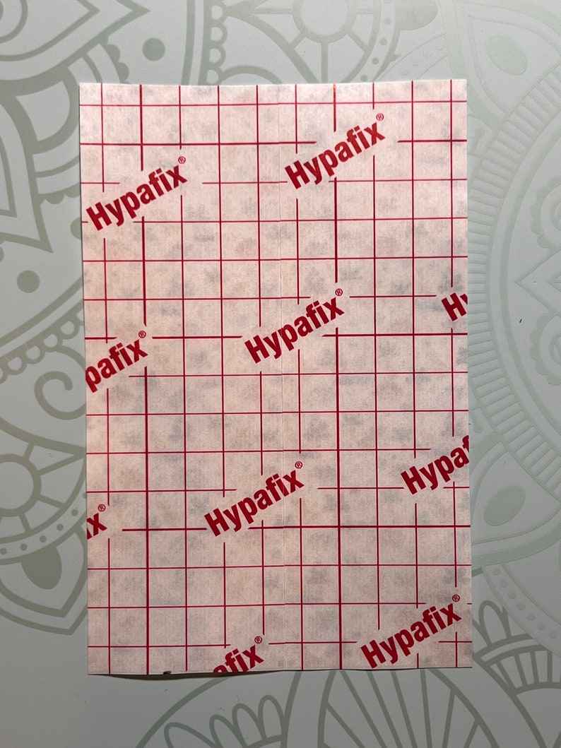 Op de afbeelding: Witte plakband met een rasterpatroon en de tekst "Hypafix" die overal herhaald wordt.