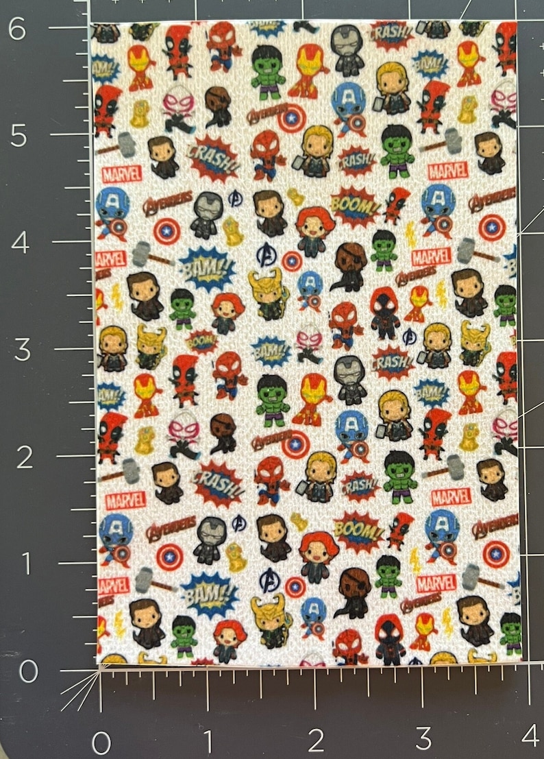 Op de afbeelding: Witte stof met een herhalend patroon van kleine cartoon Marvel-personages, waaronder Iron Man, Captain America, Thor, Hulk, Spider-Man en Deadpool. De stof bevat ook de woorden "Marvel", "Avengers", "Boom", "Crash" en "Bam!" in een comic-stijl lettertype.