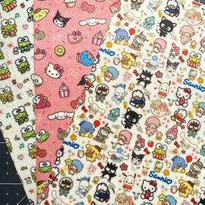 May include: Three sheets of faux leather fabric featuring Sanrio character prints. One sheet has a white background with green frogs, another is pink with Hello Kitty and sweets, and the third is white with a dense pattern of various Sanrio characters.
