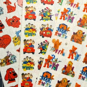 May include: Three sheets of stickers featuring colorful illustrations of Clifford the Big Red Dog, The Wiggles in their car, and characters from Bear in the Big Blue House. The stickers are arranged in rows on white paper backing with a textured surface.