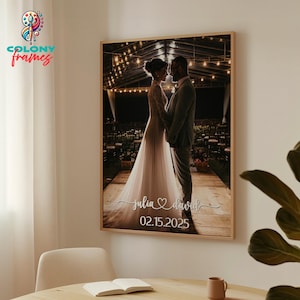 May include: A black and white photo of a bride and groom standing under a canopy of lights. The bride is wearing a white wedding dress and the groom is wearing a suit. The photo is framed in a wooden frame and has the text "julia ♡ david 02.15.2025" printed below the image.