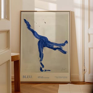 May include: Framed art print featuring a bold blue abstract figure against a neutral background. The artwork includes the word "BLEU" at the bottom left, with additional text below. The frame is a light wood color, and the artwork is displayed in a room with a door and a small wooden stool.