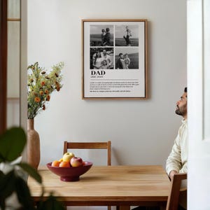 May include: A framed print with a wooden frame featuring four black and white family photos. The word "DAD" is printed below the photos, along with a definition. The print hangs on a white wall above a wooden table with a maroon bowl of fruit.
