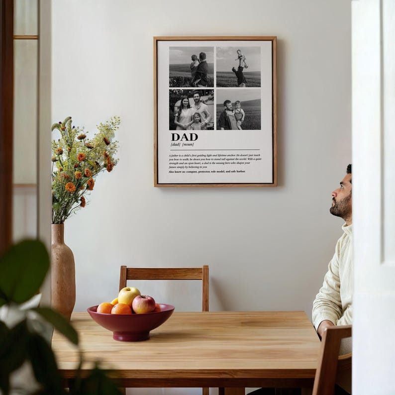 May include: A framed print with a wooden frame featuring four black and white family photos. The word "DAD" is printed below the photos, along with a definition. The print hangs on a white wall above a wooden table with a maroon bowl of fruit.