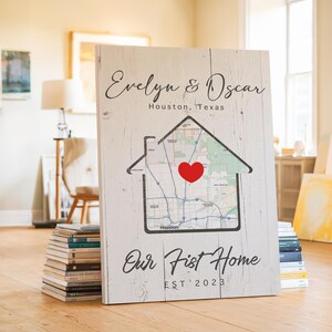 May include: A white wooden sign with the text "Evelyn & Oscar" and "Houston, Texas" in black script. A house-shaped outline contains a map with a red heart. The text "Our First Home EST 2023" is below the house graphic.