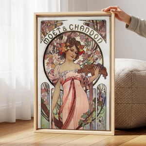 May include: A framed art print featuring a vintage advertisement for Moët & Chandon champagne. The artwork depicts a woman in a pink dress holding grapes, with the brand name in gold lettering. The print has a wooden frame.