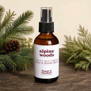 May include: An amber glass bottle of Alpine Woods linen and room spray with a black spray nozzle. The white label features the text Alpine Woods, balsam fir, crisp air, cedarwood, Linen & Room Spray, flower & meadow, and Net Wt. 3.4oz (100ml).