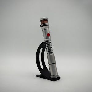 Op de afbeelding: Een zilverkleurig metalen lichtzwaardhandvat met een geribbelde grip, een koperkleurige accentring bij de zender en een opvallende rode ronde knop aan de zijkant, rechtopstaand gepresenteerd op een gebogen zwarte kunststof standaard.