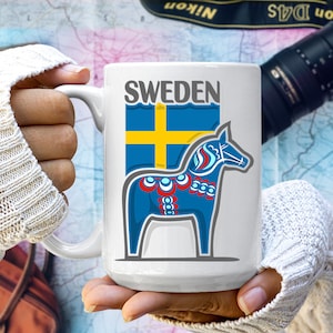 May include: White ceramic mug with a blue and yellow Swedish flag and a blue and yellow Dala horse design. The mug says "SWEDEN" in gray text.