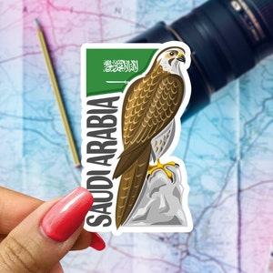 May include: A sticker featuring a brown and white falcon perched on a rock, with the text "SAUDI ARABIA" in white. The sticker has a green border with Arabic text and a white sword. The background includes a map and a camera lens.