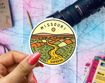 May include: A round sticker with a yellow background and a black outline. The sticker features a stylized landscape with a winding river and the words "MISSOURI SALUS POPULI SUPREMA LEX ESTO" in a black circle.