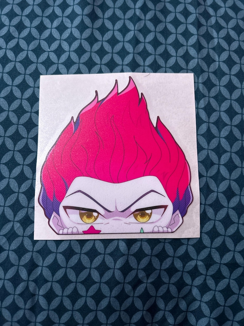 Puede incluir: Una pegatina con un personaje de dibujos animados con cabello rojo, ojos amarillos y una estrella rosa en la mejilla. El personaje est&aacute; mirando al espectador mientras se asoma por encima de una superficie.