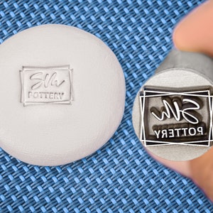 Op de afbeelding: Een close-up van een metalen stempel met de tekst "Silh Pottery" in een rechthoekig frame. De stempel wordt vastgehouden door een hand, en de tekst is omgekeerd, alsof deze in klei moet worden gedrukt.