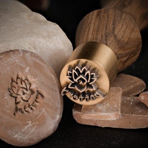Könnte beinhalten: Ein Holzgriff mit einem Messing-Stempelkopf mit einem Lotusblumen-Design. Der Stempel wird verwendet, um Abdrücke in Ton oder anderen weichen Materialien zu erstellen. Der Text "Pottery" ist in den Stempelkopf graviert.