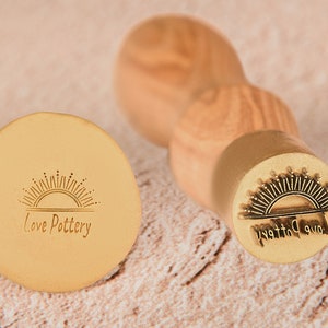 Peut inclure: Un tampon en métal doré avec une poignée en bois. Le tampon présente un motif de soleil levant et le texte "Love Pottery".