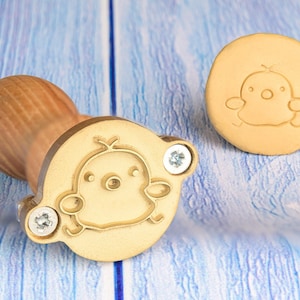 May include: A gold-toned metal stamp with a wooden handle features a chick design. The stamp is used for creating impressions on wax or clay.