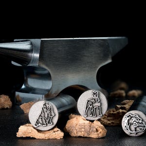 Könnte beinhalten: Drei Stempel aus Metall mit Designs von Hunderassen. Die Stempel befinden sich auf einer Holzoberfläche neben einem Amboss aus Metall.