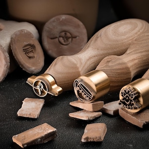 Può includere: Quattro timbri in argilla con manici in legno e teste in ottone. I timbri presentano diversi disegni, tra cui un fiore, un vortice, un cuore e un motivo geometrico. I timbri sono su una superficie scura con pezzi di argilla rotta.