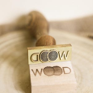 Op de afbeelding: Een houten stempel met een messing bovenkant. De messing bovenkant heeft het woord "WOOD" met een gestileerd boomringontwerp. De houten basis heeft het woord "WOOD" met een gestileerd boomringontwerp.