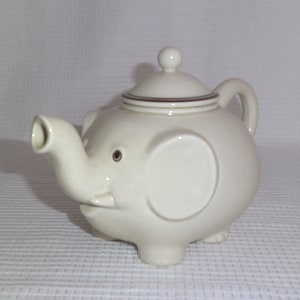 May include: A white ceramic teapot shaped like an elephant with a brown stripe around the lid. The teapot has a spout shaped like an elephant's trunk and a handle on the side.